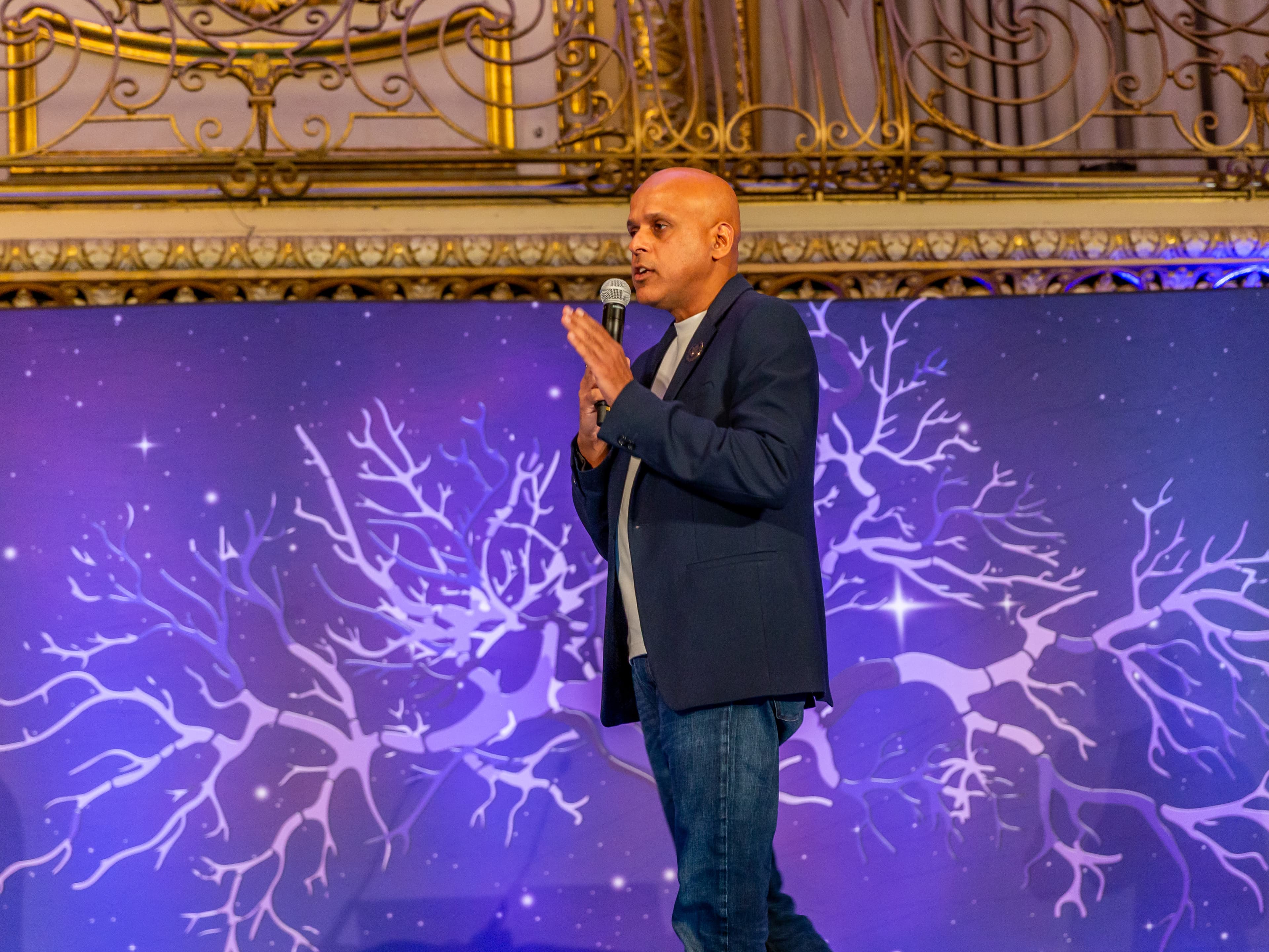 Manuj Aggarwal speaking with a microphone against a purple backdrop.